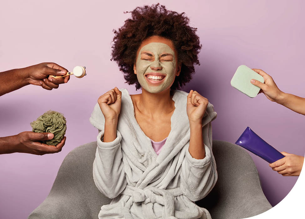 Skin care and beauty concept. Photo of overjoyed Afro American woman in bathrobe clenches fists with happiness, applies facial clay mask on face, rejoices having perfect skin. Hands with cosmetics